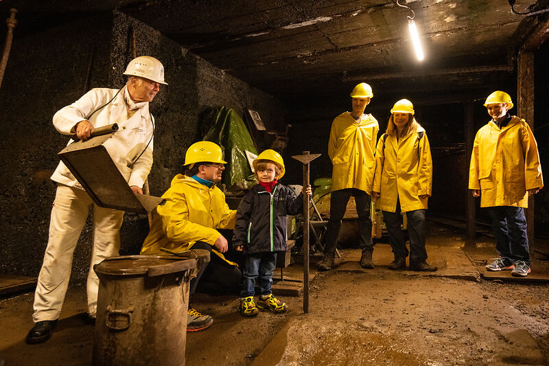 Mehrere Besucher in gelben Mänteln und gelben Helmen in einem dunklen Raum. Ein Kind steht in der Mitte und hält ein Werkzeug nach oben.
