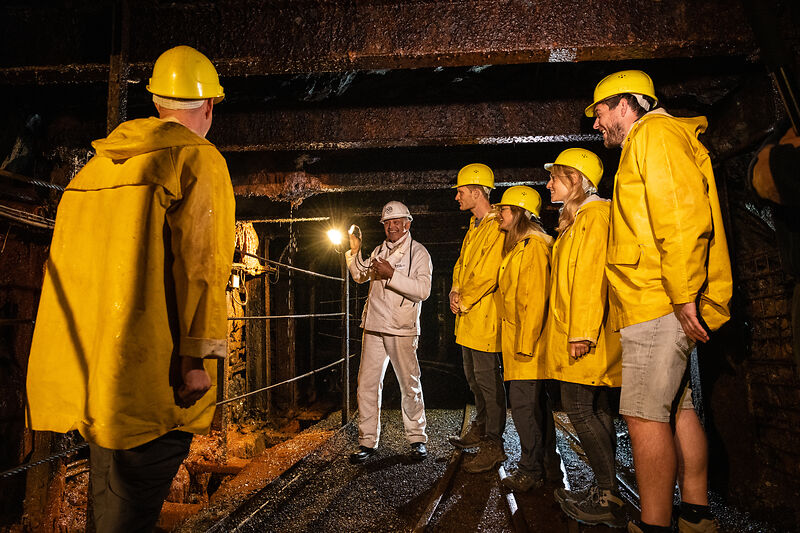 Mehrere Besucher in gelben Mänteln und gelben Helmen besichtigen die Grube, ein Mann in weiß gekleidet spricht mit den Besuchern
