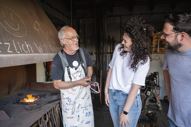 Ein Schmied unterhält sich mit einem Pärchen, alle Personen lachen